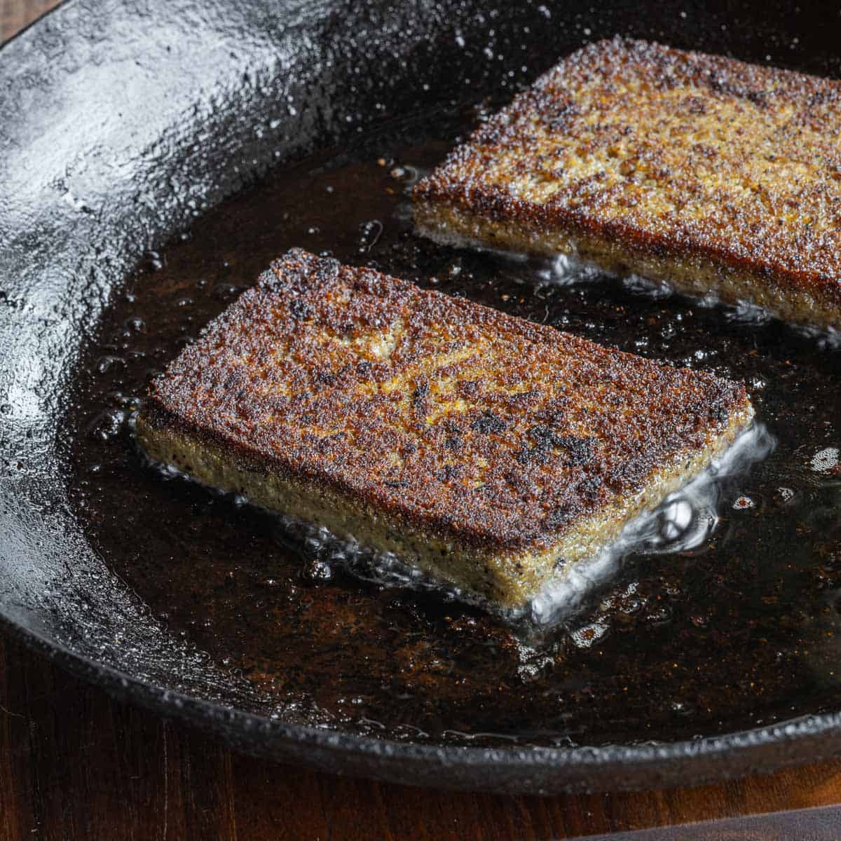 Two slices of golden brown scrapple cooking in a carbon steel pan. 