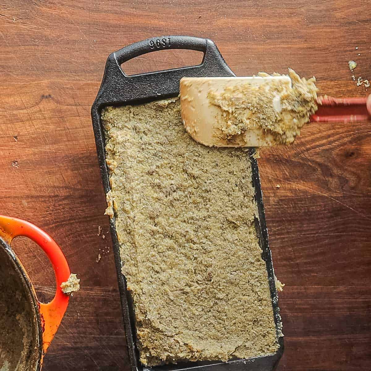 Spreading cooked scrapple into a greased cast iron loaf pan with a spatula. 