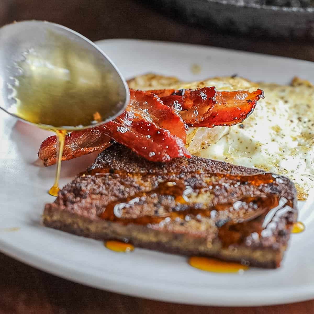 A spoon drizzling maple syrup over a plate of scrapple slices, eggs and bacon. 