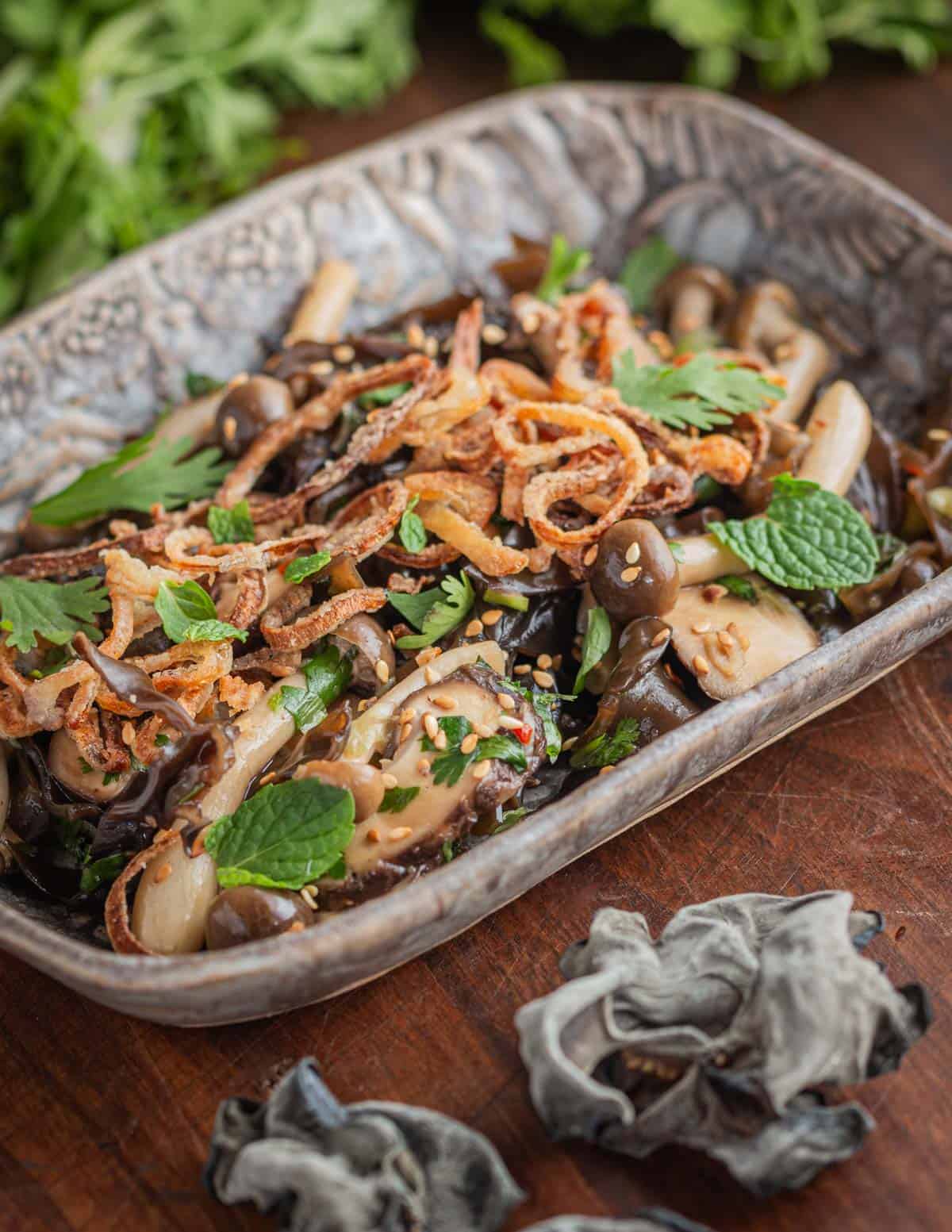 A semi traditional dried wood ear mushroom salad garnished with fried shallots and herbs on a ceramic serving dish.
