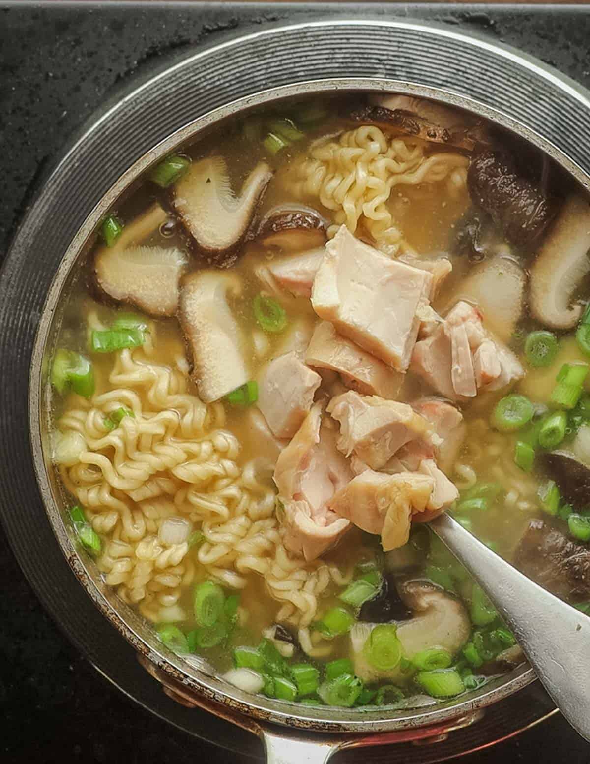 Adding cooked chicken and scallions to a bowl of ramen noodles in a pot. 