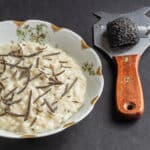 A bowl of black truffle risotto topped with chopped truffles next to a truffle grater with a black truffle on it.