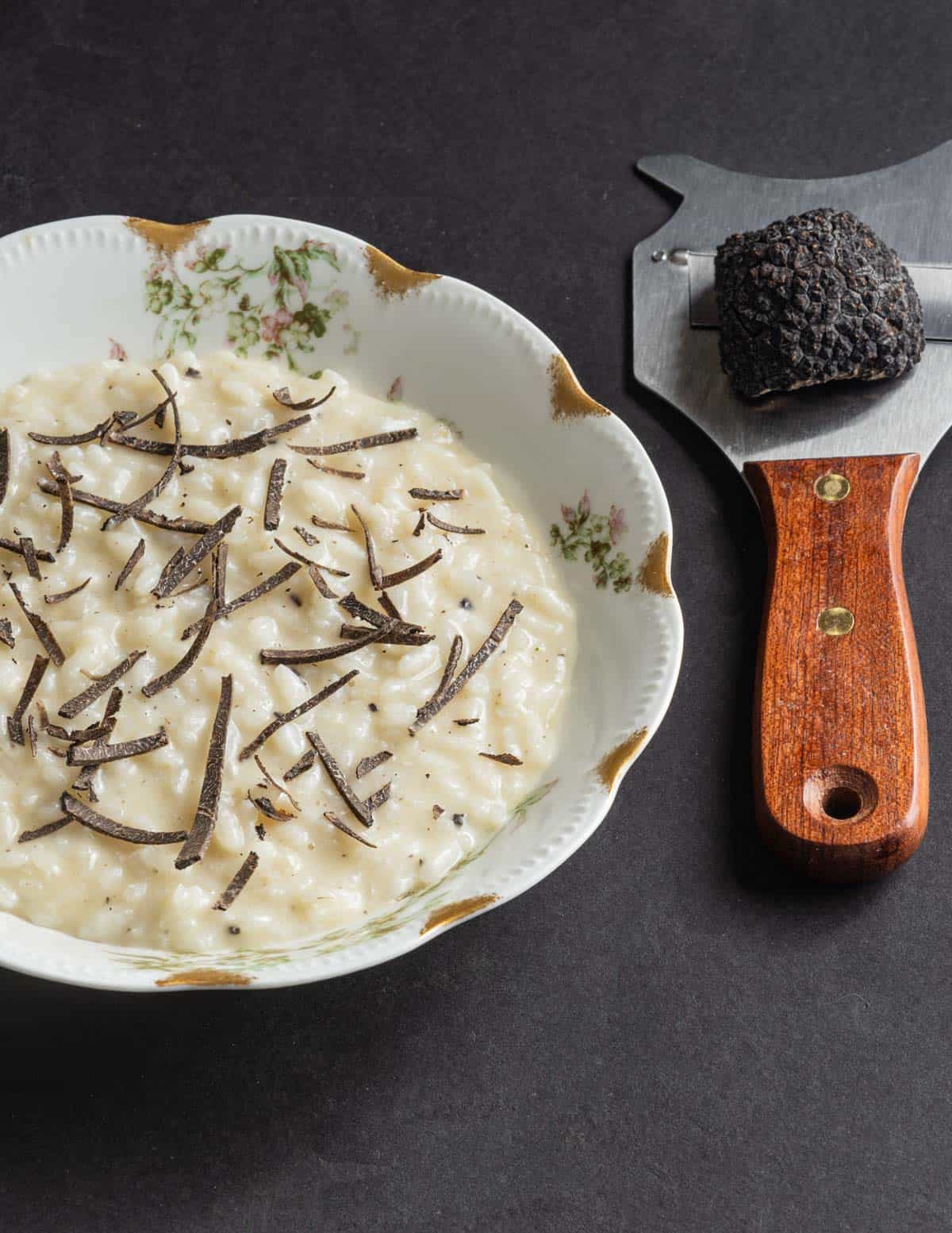 A bowl of black truffle risotto topped with shaved black truffles next to a truffle slicer with a truffle on top. 