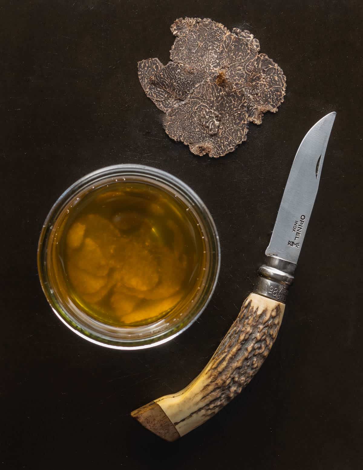 A knife next to sliced black perigord truffles and a jar of homemade black truffle oil.