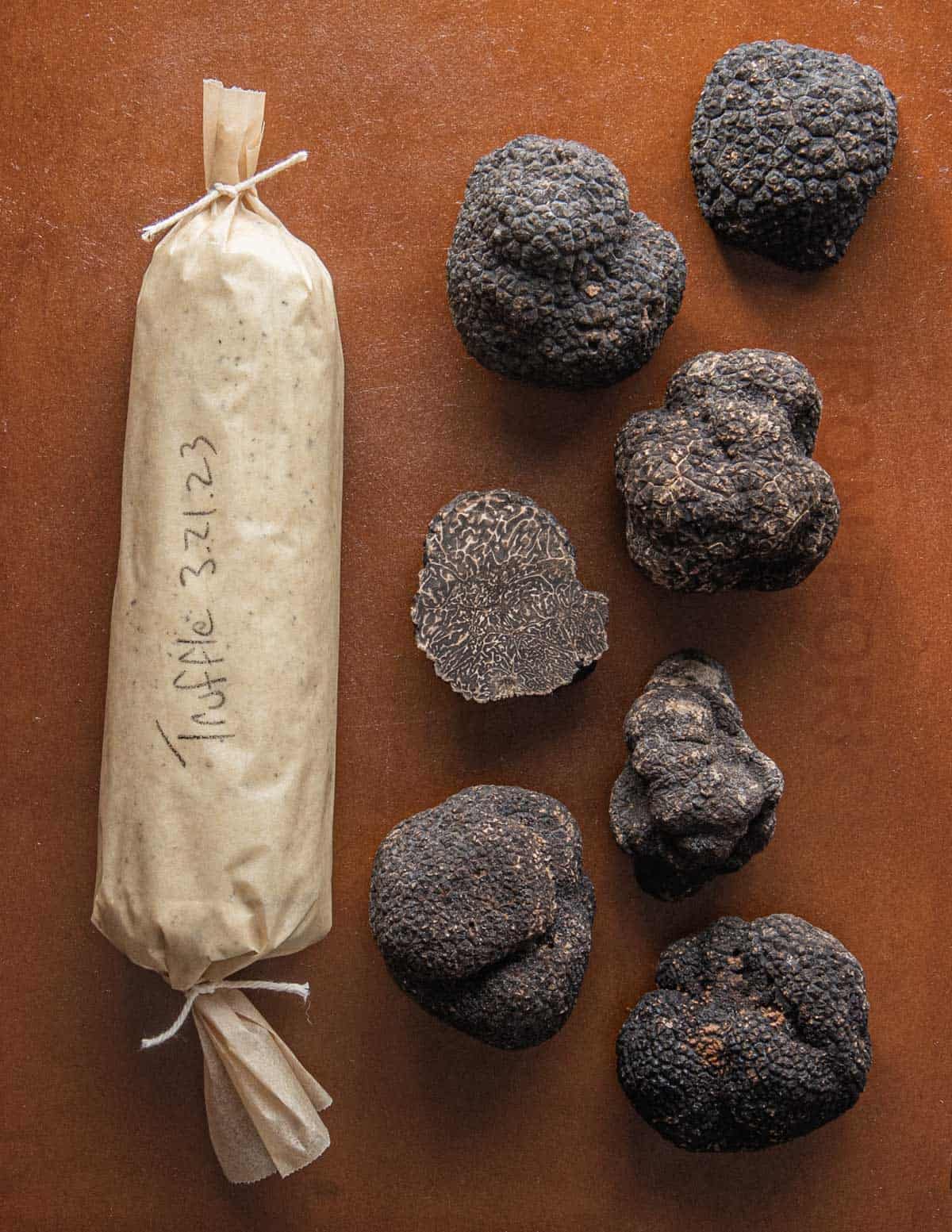 A log of black truffle butter wrapped in parchment next to many fresh black Perigord truffles. 