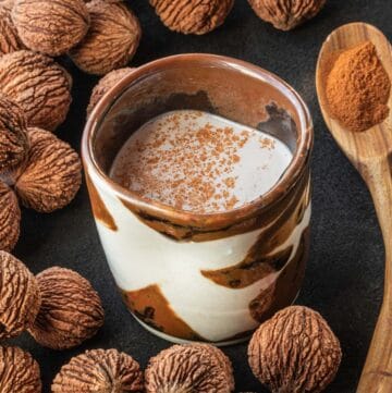 A warm glass of black walnut milk pictured next to dried black walnuts in the shell and a spoonful of cinnamon.