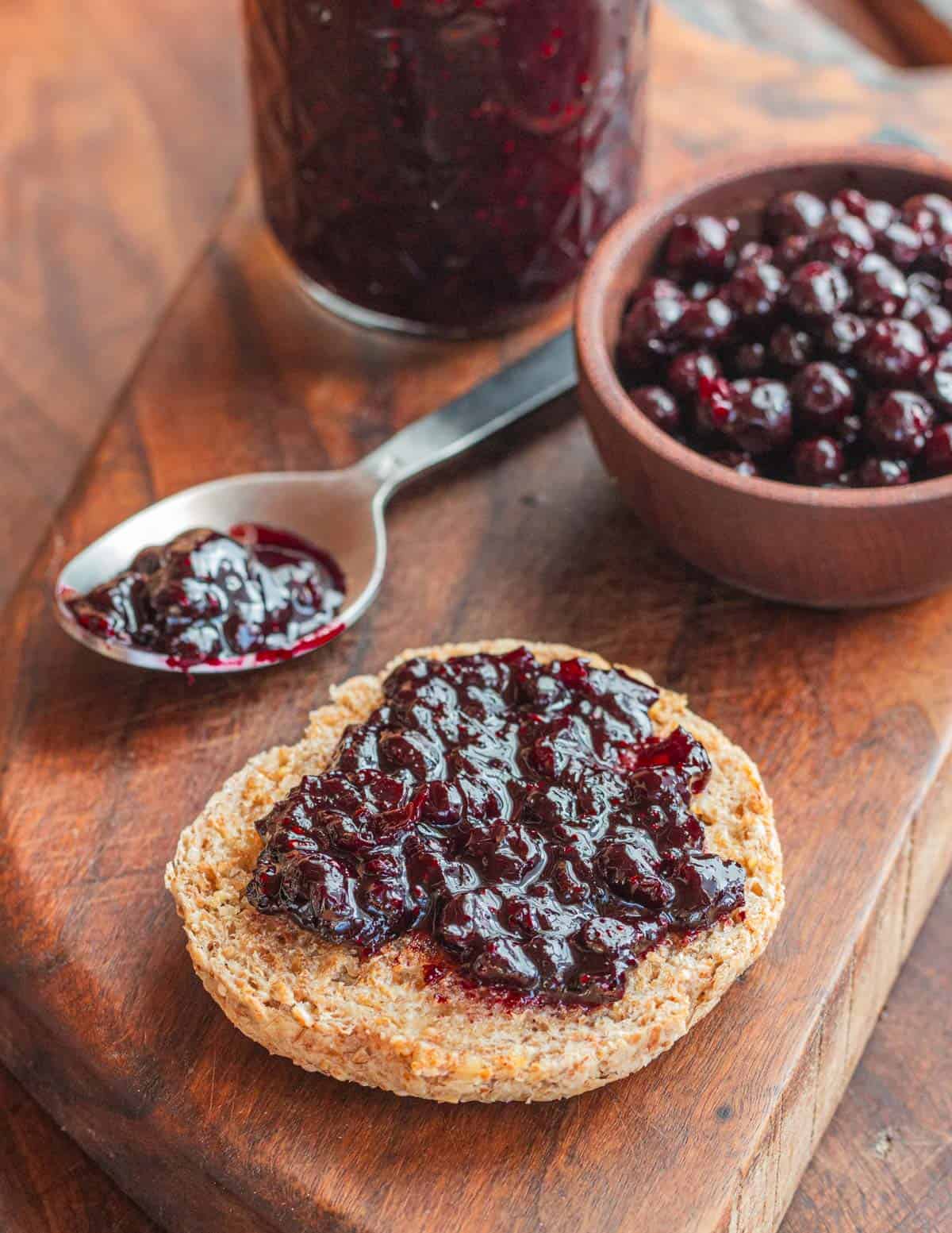 A buttered English muffin spread with black currant jam next to a bowl of fresh black currants and jar of jam.