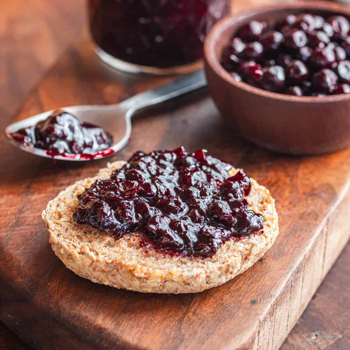 A buttered English muffin spread with freshly cooked black currant preserves on a board next to a jar of currant jam and a bowl of fresh black currants. 