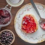 An English muffin buttered and spread with chokecherry jelly on a china plate next to a bowl of cherries and a jar of jelly.