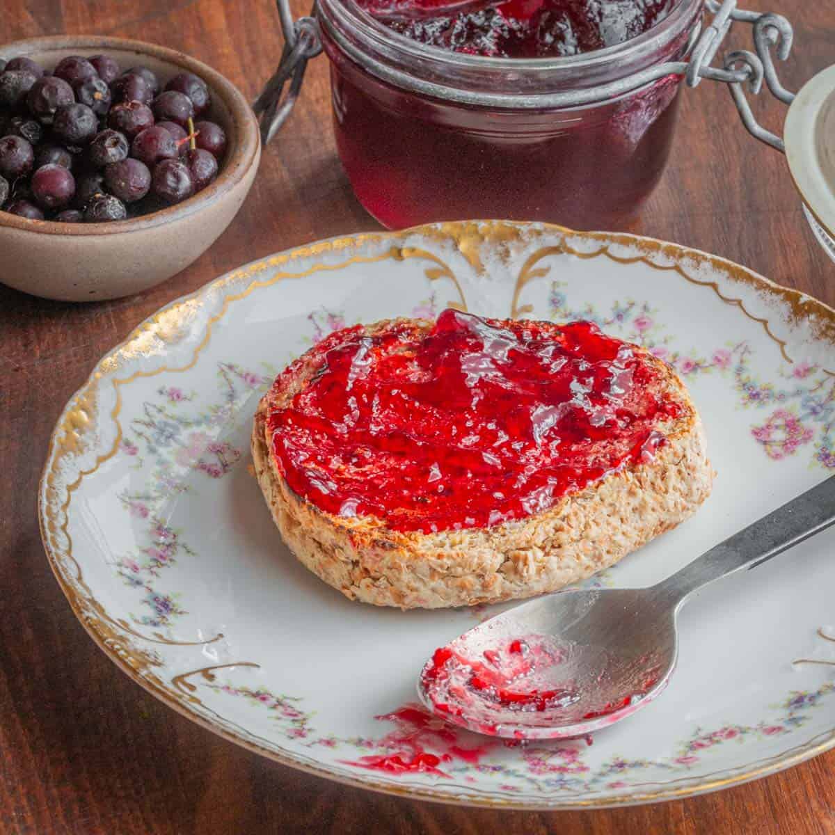 An English muffin spread with chokecherry jelly next to a jar of jelly and a bowl of wild cherries.