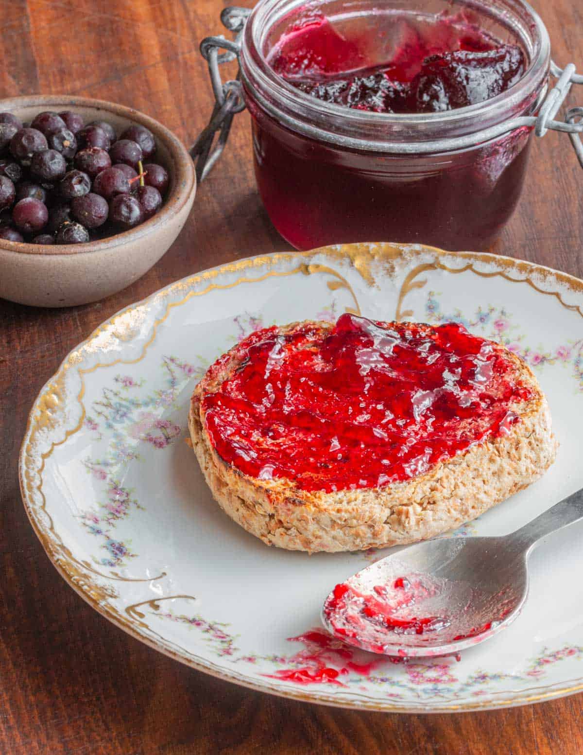 Choke cherry jelly spread on an English muffin next to a jar of jelly and a bowl of wild cherries.