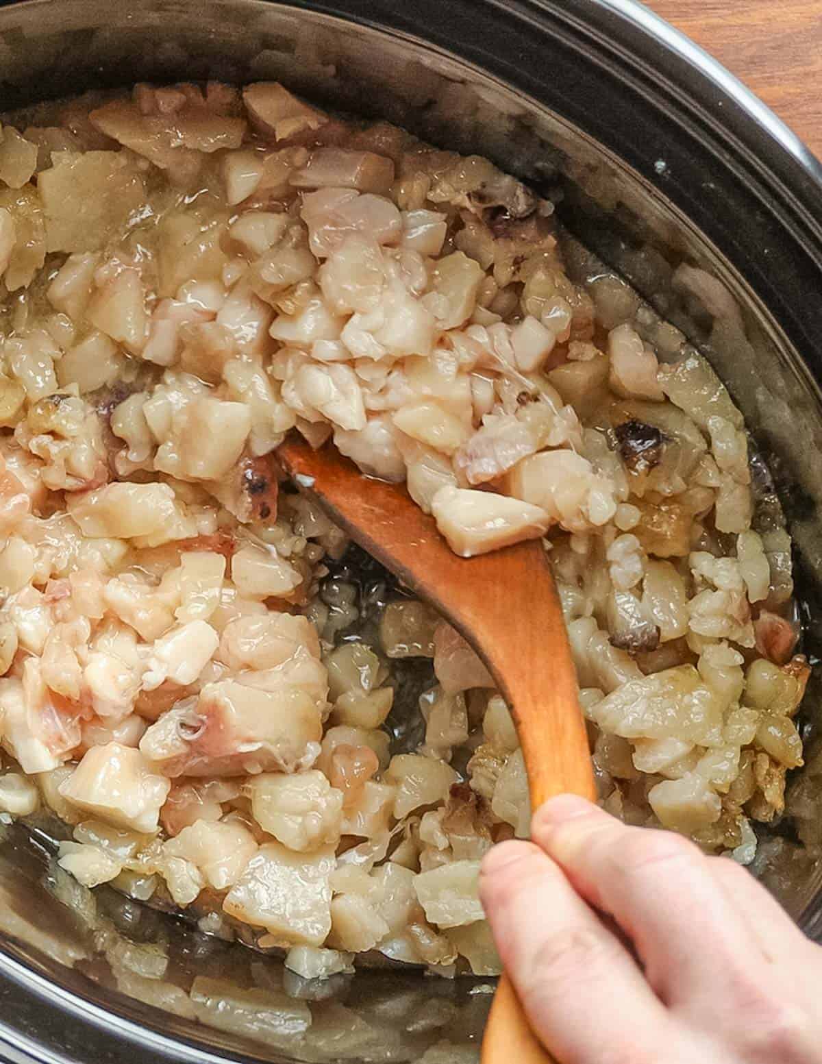Stirring a slow cooker filled with large pieces of beef fat rendering into tallow. 