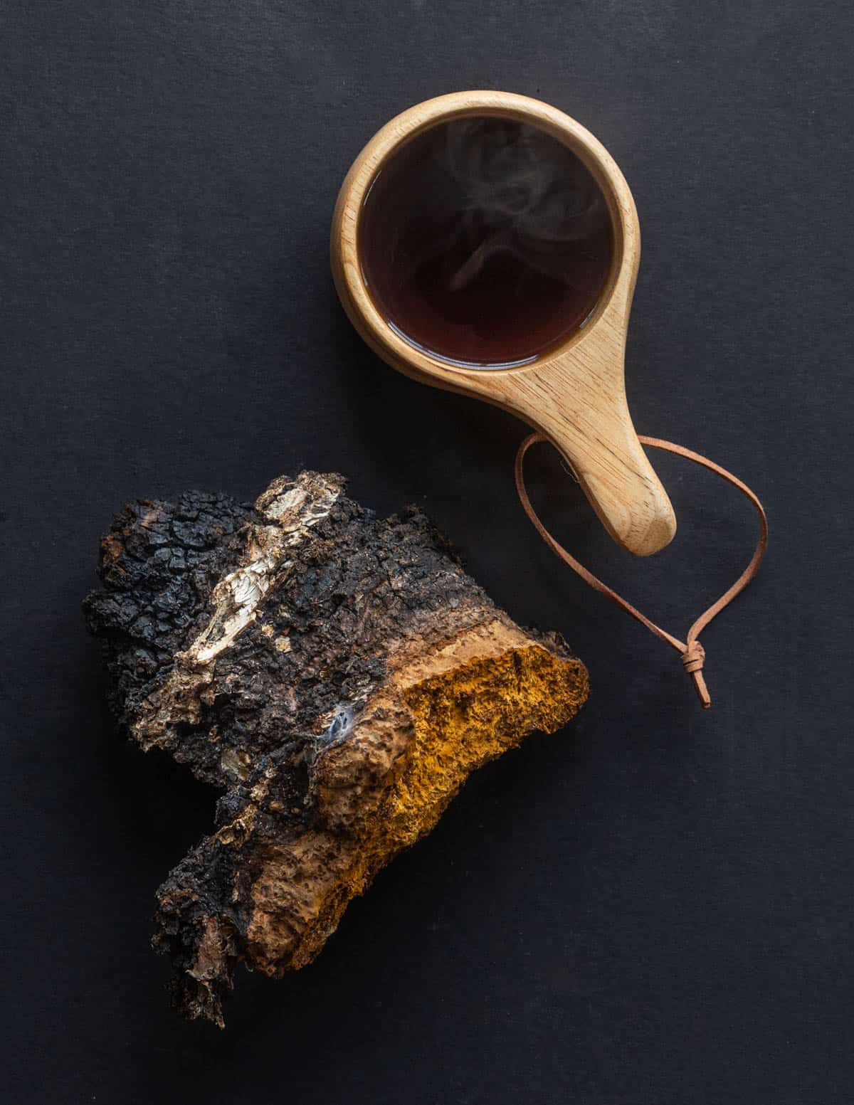 A cup of dark chaga tea on a black background next to a whole fresh chaga mushroom. 