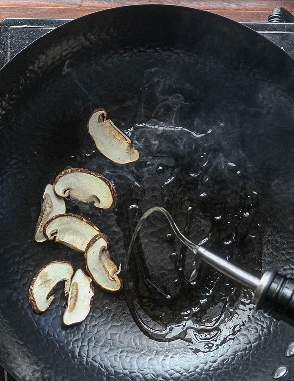 Sliced shiitake mushrooms cooking in a wok with oil being added after sauteeing. 