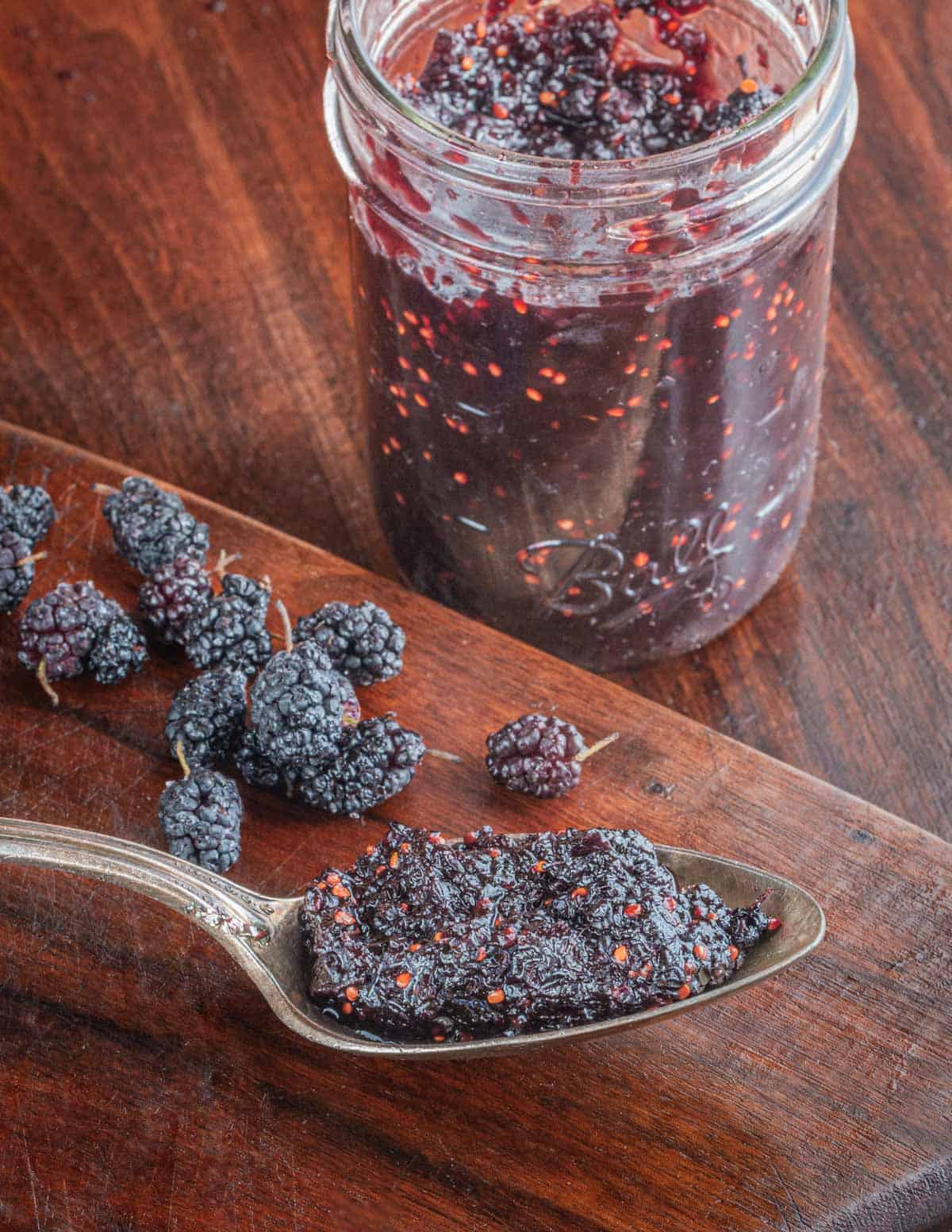 A jar of homemade mulberry jam next to a spoonful of jam and fresh mulberries. 