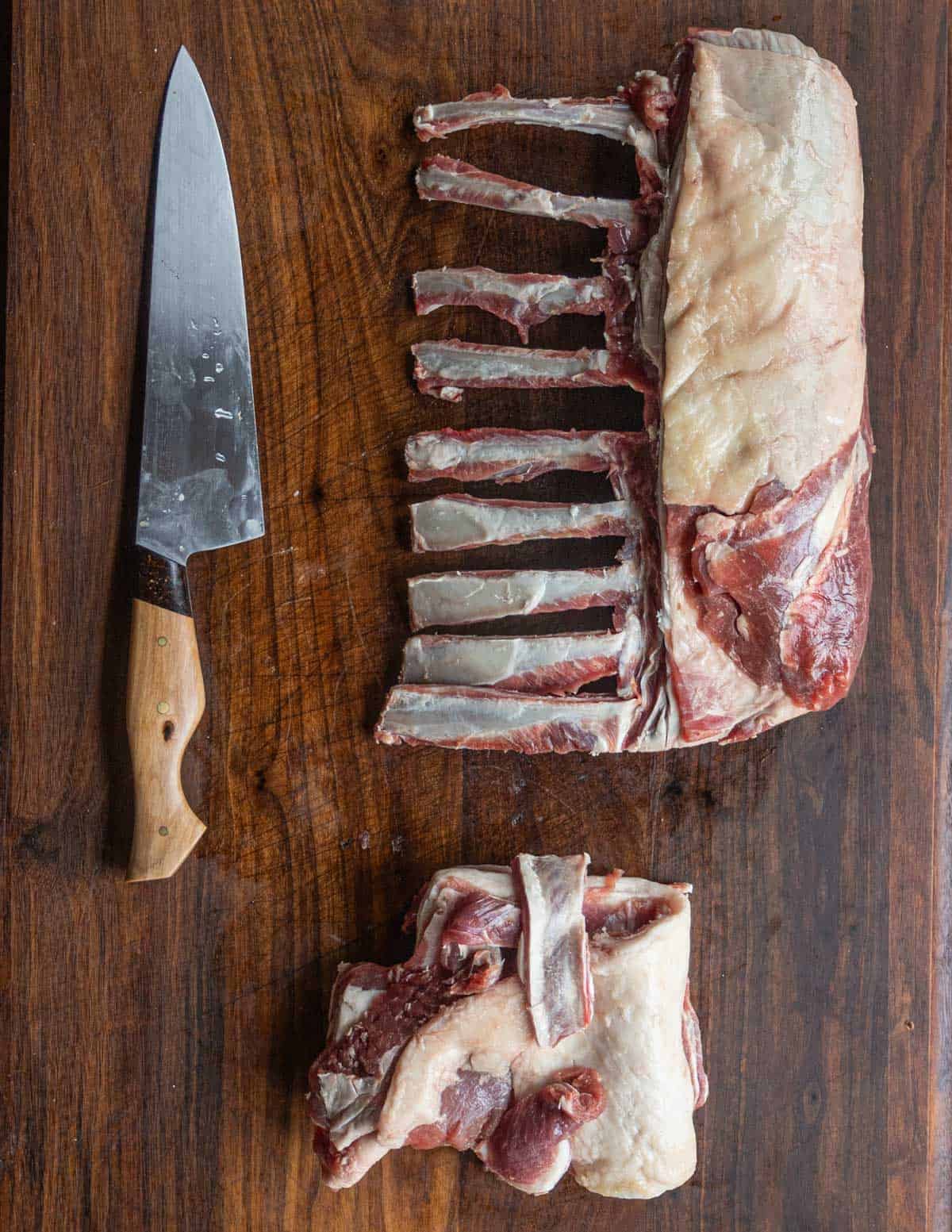 A rack of lamb trimmed and frenched showing a pile of trim and a cleaned rack of lamb next to a knife on a cutting board. 