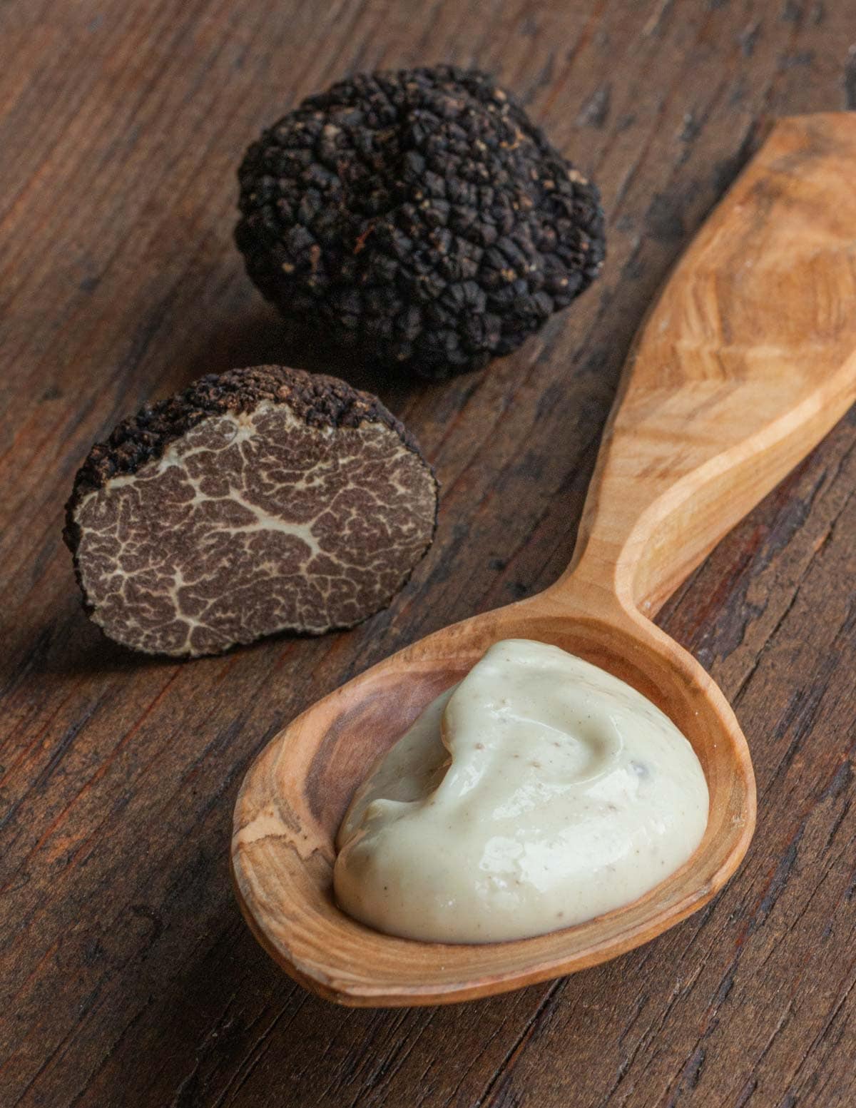 A spoon filled with garlic black truffle aioli next to a fresh black truffle on a cutting board.
