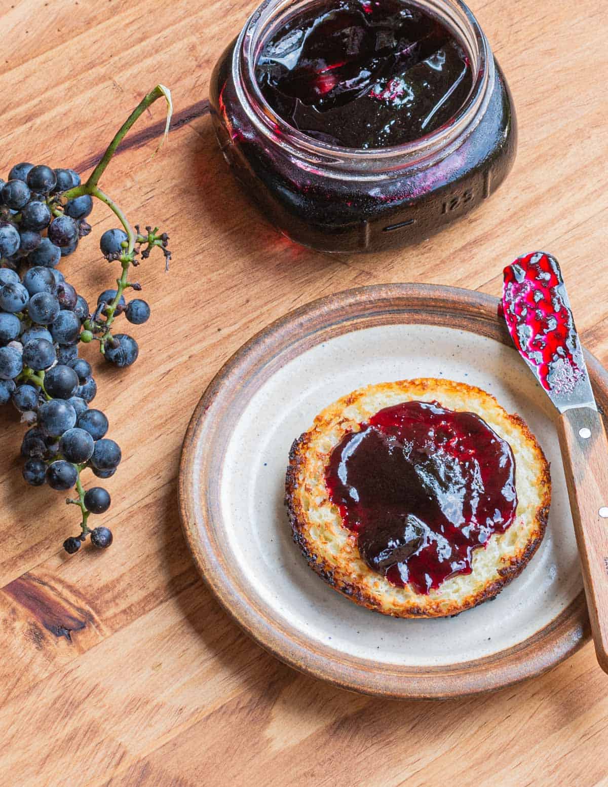 An English muffin with wild grape jelly next to a jar of jelly and a cluster of wild grapes. 
