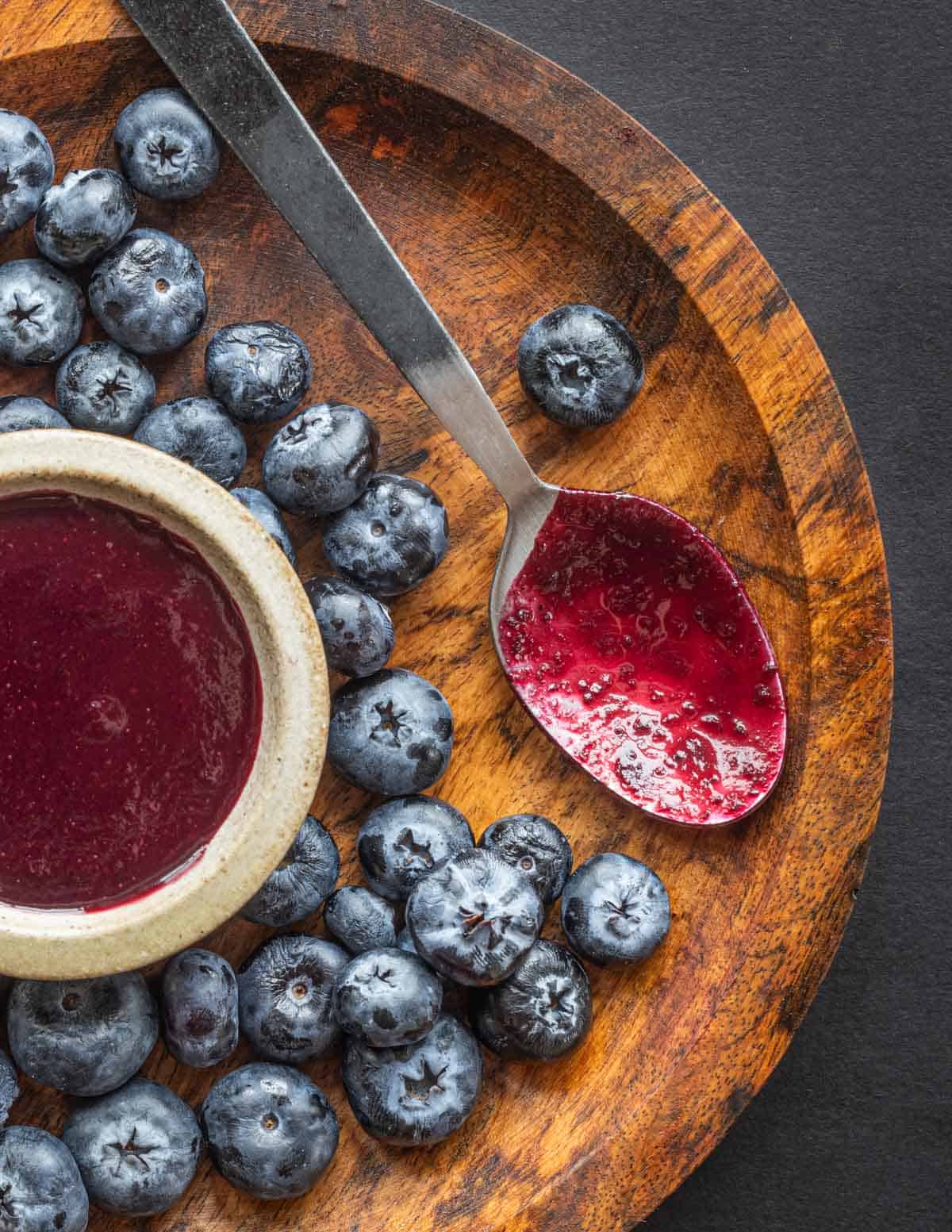 A close up image of a spoon purple with wild blueberry barbecue sauce on a wooden plate next to a bowl of wild blueberry barbecue sauce and fresh blueberries.