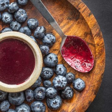 A close up image of a spoon purple with wild blueberry barbecue sauce on a wooden plate next to a bowl of wild blueberry barbecue sauce and fresh blueberries.