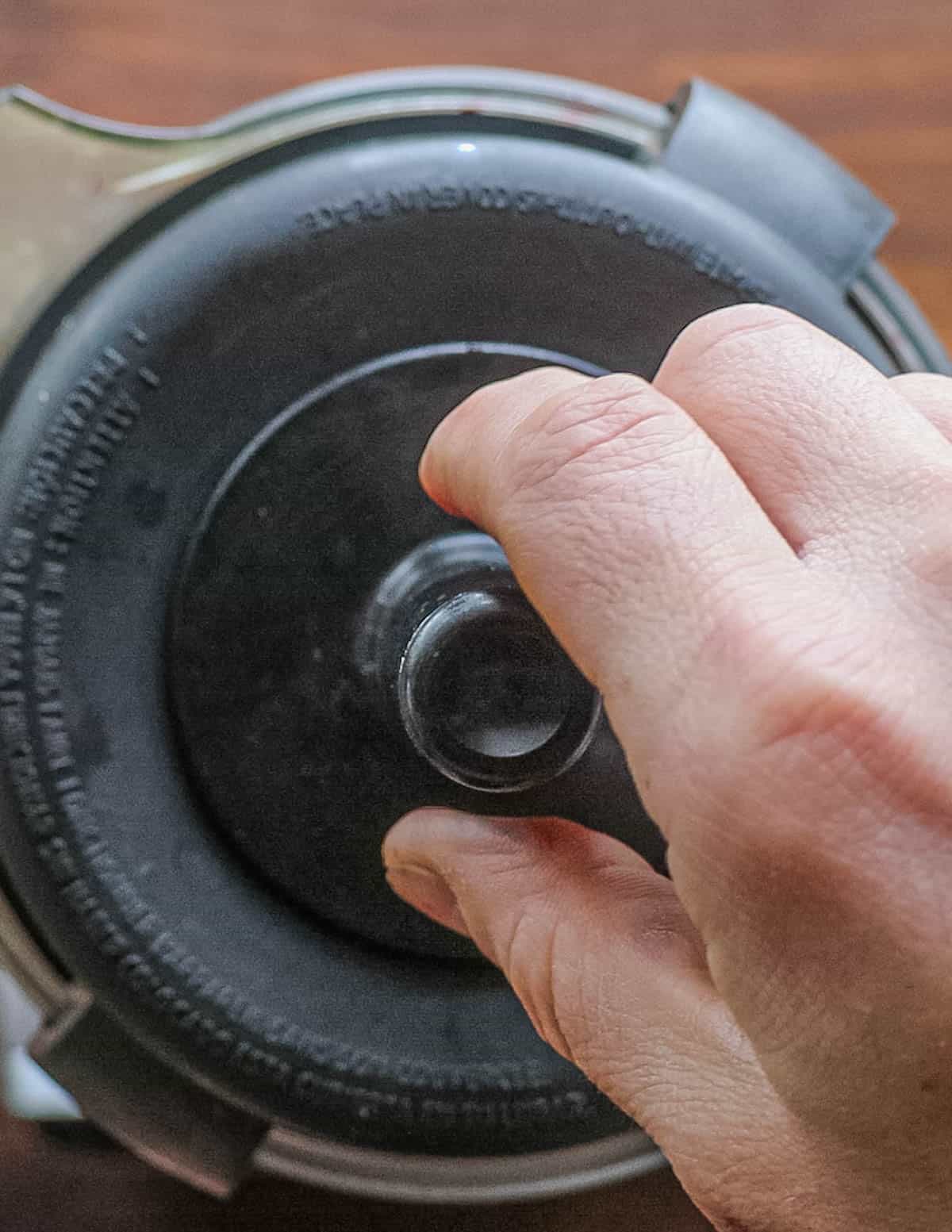 A hand holding the tamper attachment of a highspeed blender while pureeing blueberry sauce.