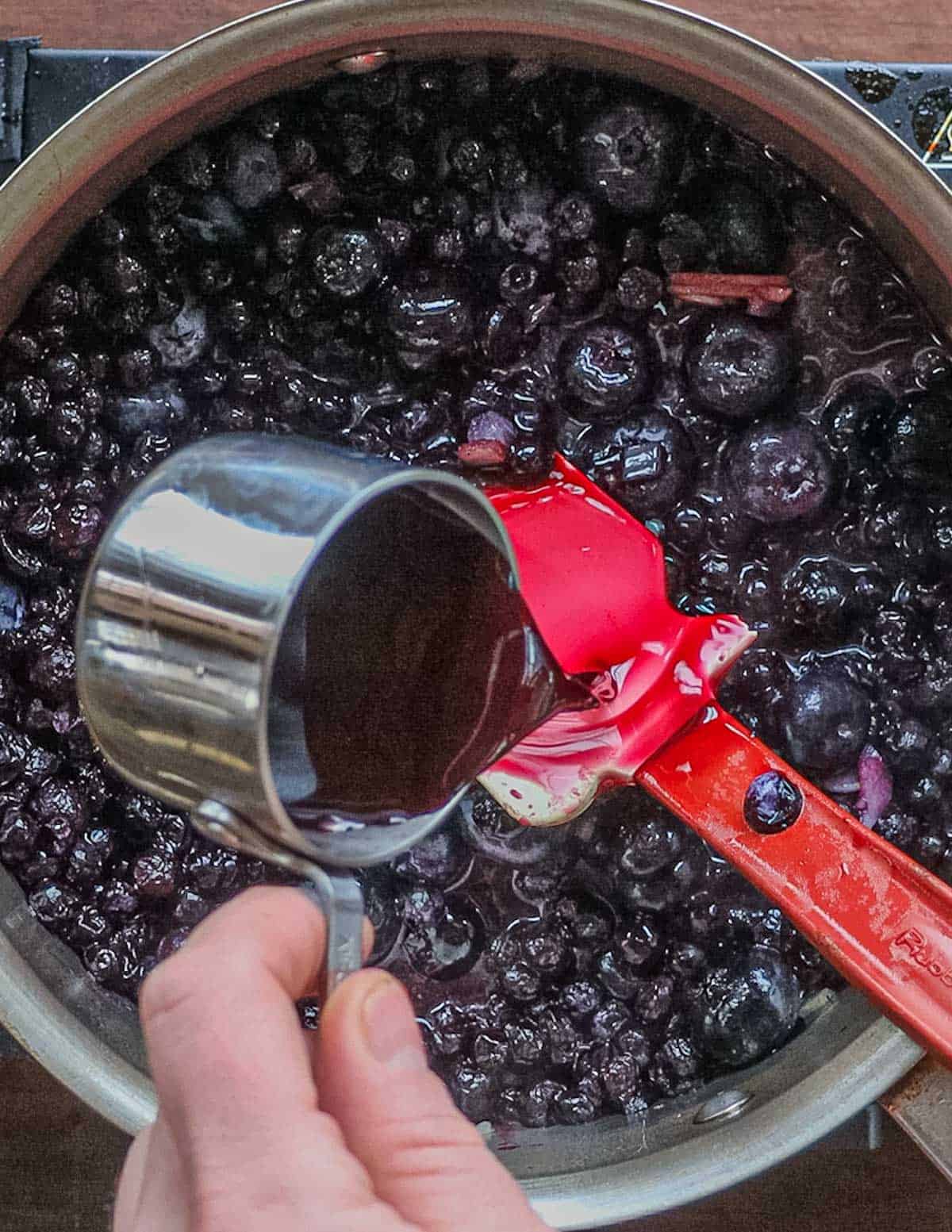Adding blueberry vinegar to a pan of cooking blueberry sauce.