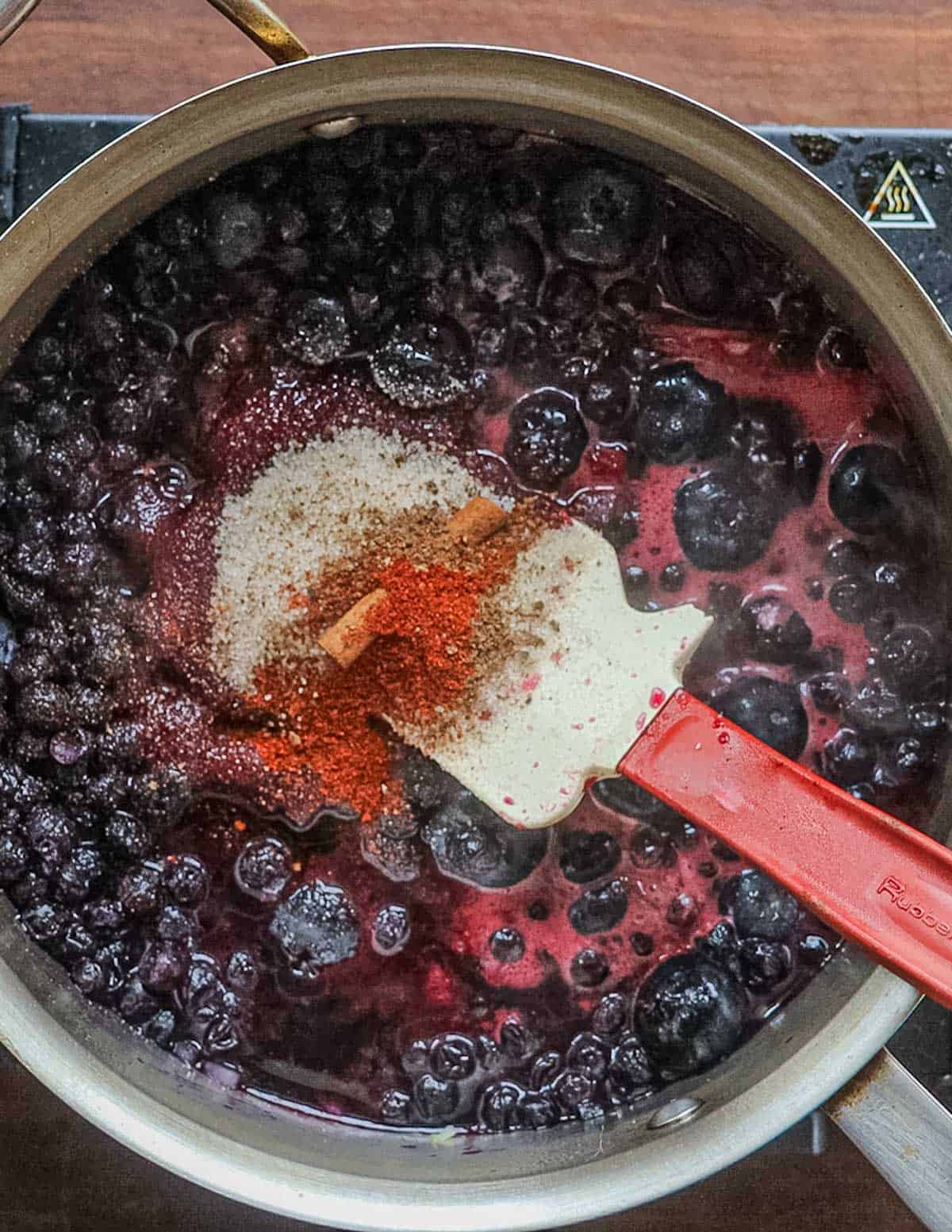 Adding ground cayenne pepper to a pan of blueberries cooking on the stove.
