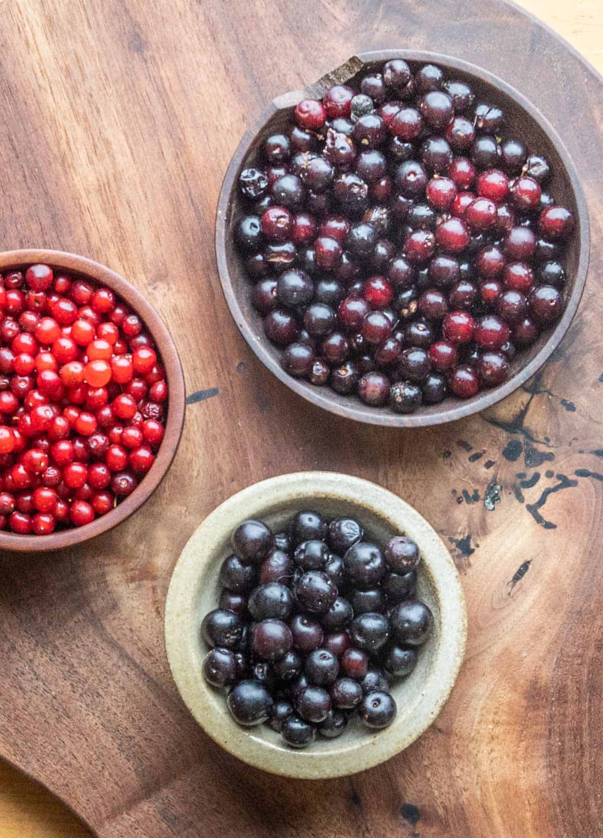 Three different types of wild cherries in bowls on a cutting board. 