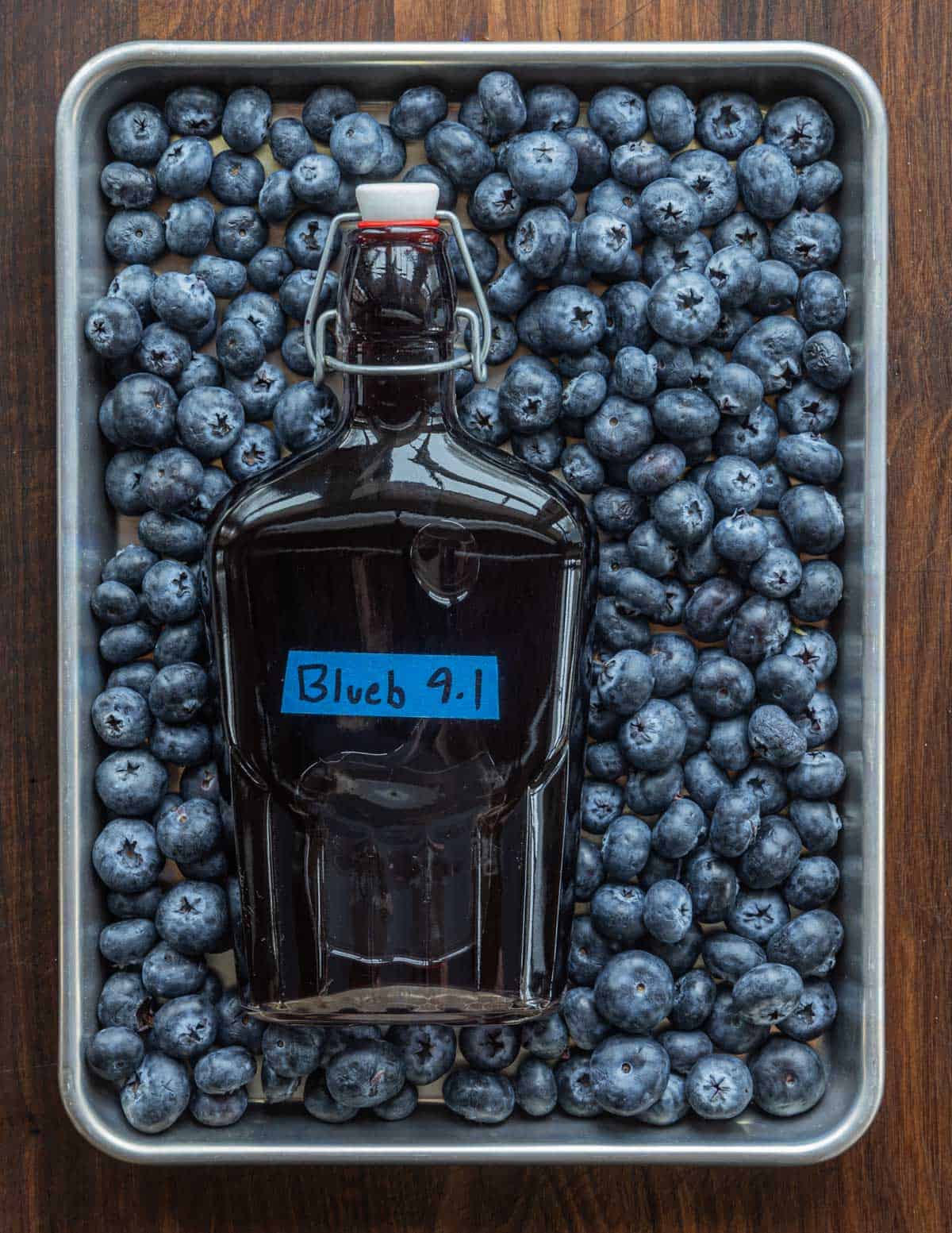 A finished jar of blueberry liqueur on a baking sheet surrounded by fresh blueberries. 