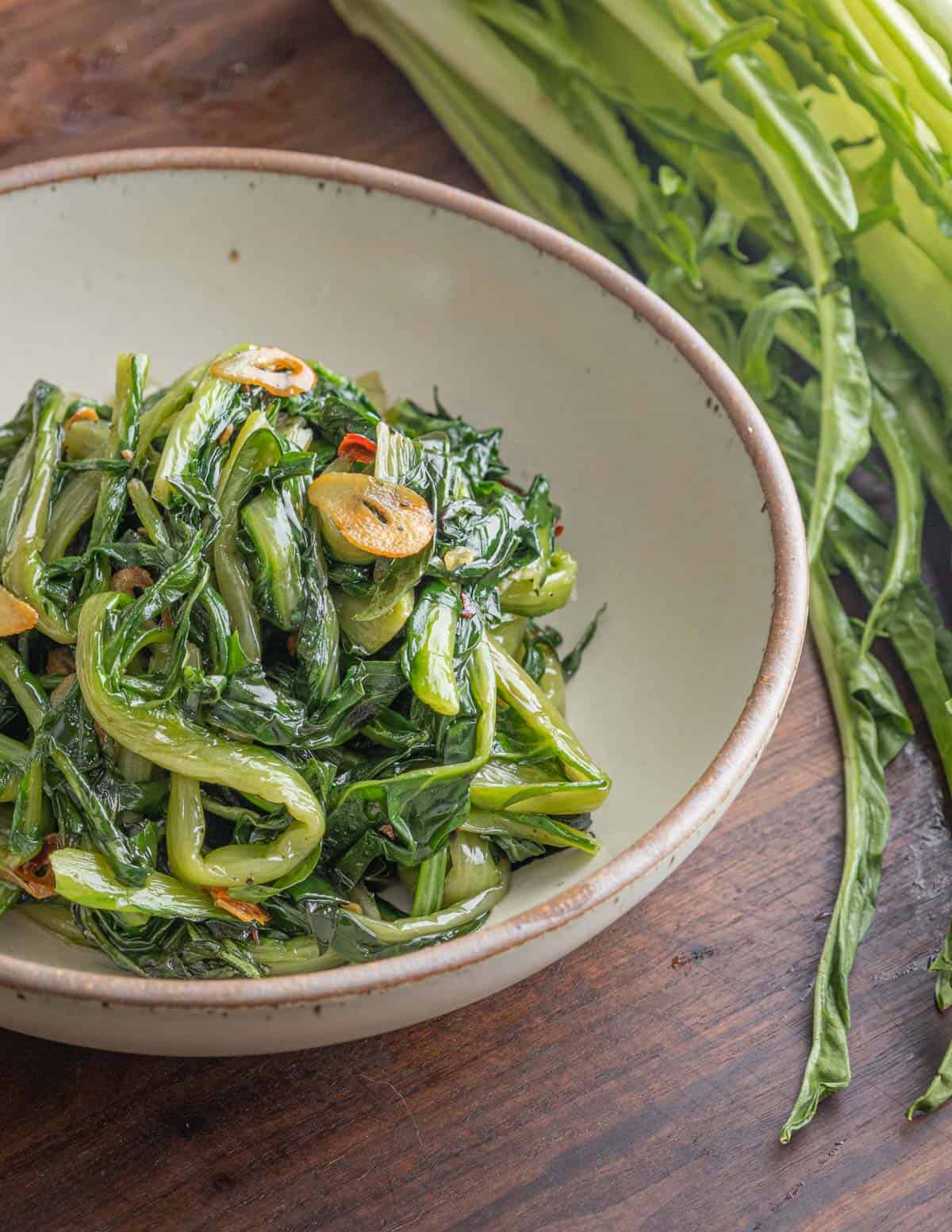 A bowl of cooked Italian puntarelle chicory greens in a bowl next to a bunch of fresh chicory greens. 