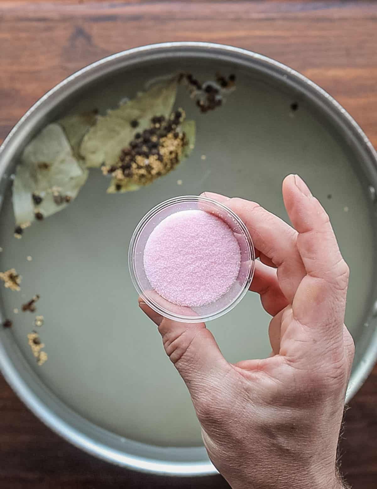 A hand holding a container of pink salt being added to a brine. 