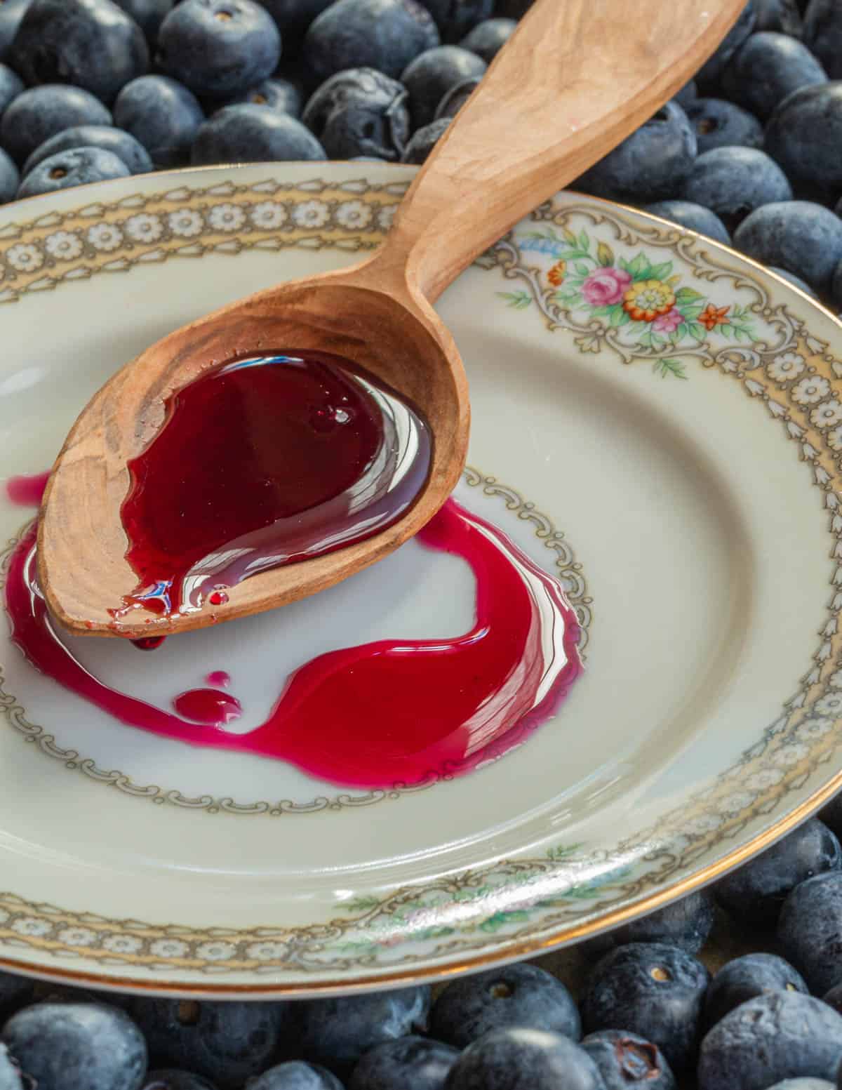 A hand-carved spoon of apple wood filled with blueberry syrup drizzled on a plate surrounded by fresh blueberries.