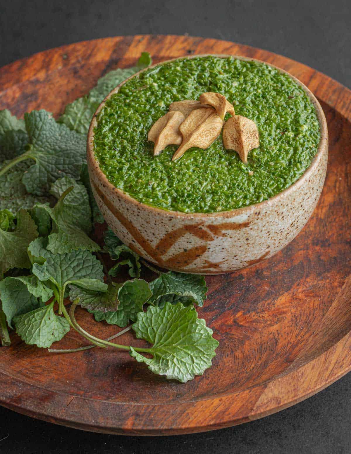 Garlic mustard pesto in a bowl surrounded by garlic mustard leaves and garnished with butternuts. 