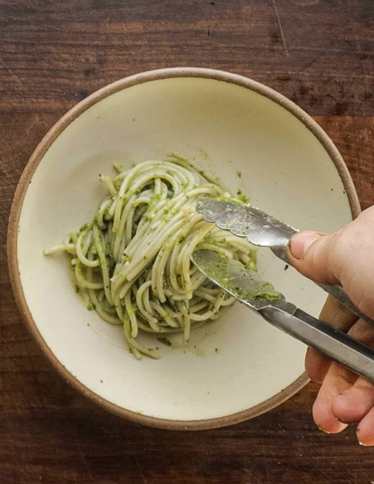 Twirling noodles with pesto into a bowl to serve using tongs. 