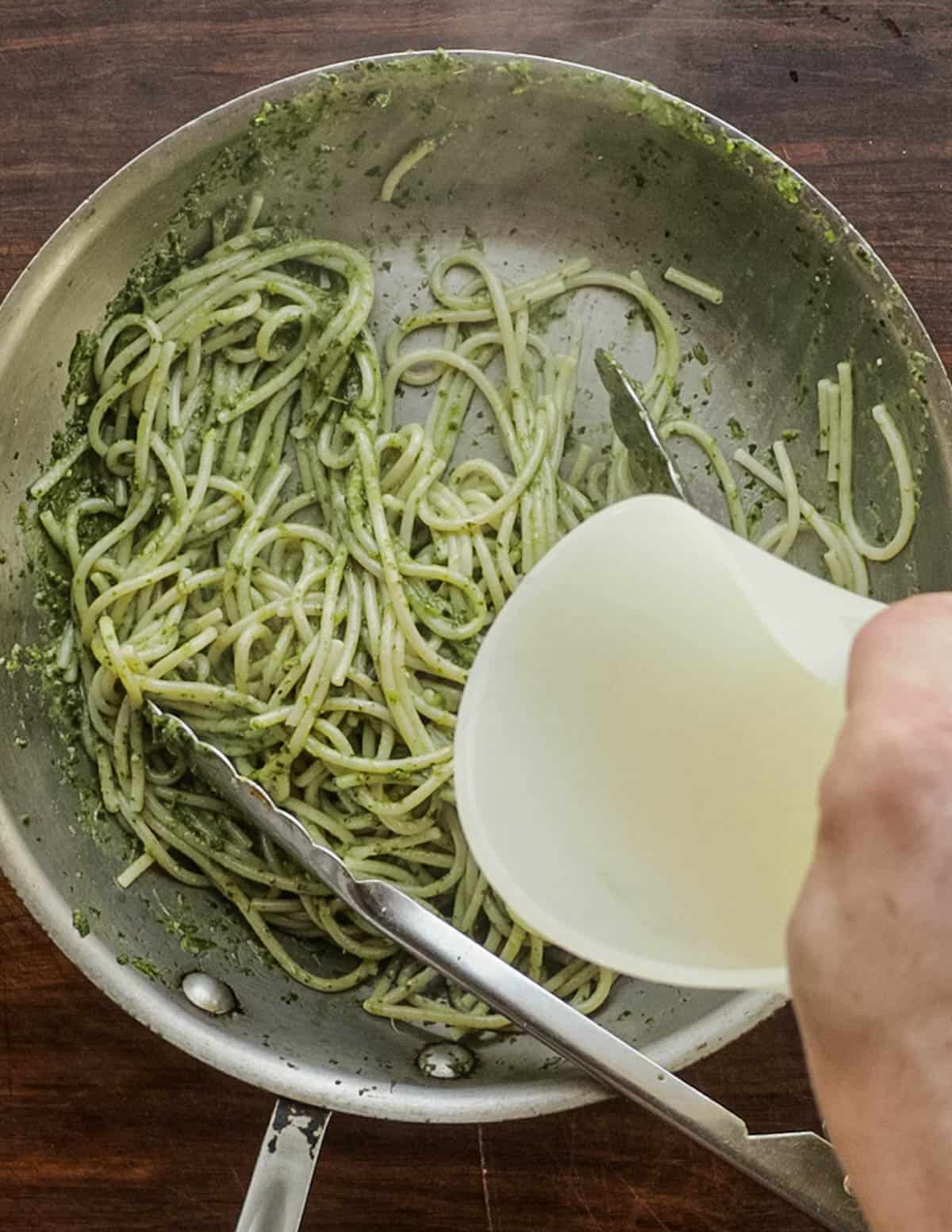Adding pasta cooking water to a pan of spaghetti with pesto. 