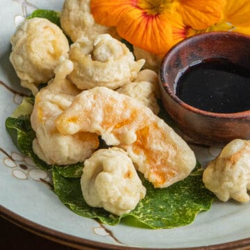 Close up image of tempura mushrooms on a plate garnished with nasturtium leaves and dipping sauce.