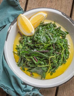 A dish of Greek horta greens seasoned with olive oil in a serving dish with fresh lemon wedges on the side. 