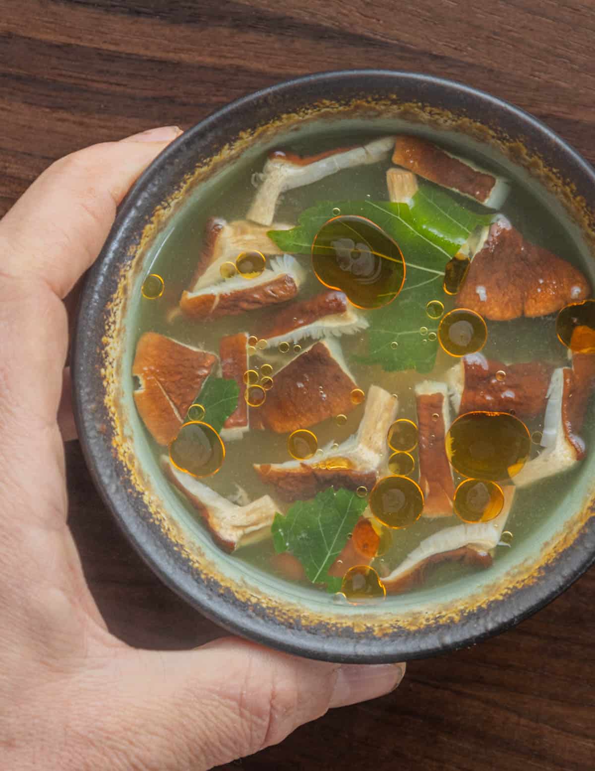A hand holding a cup of miso soup with mushrooms, tofu and mulberry leaves. 