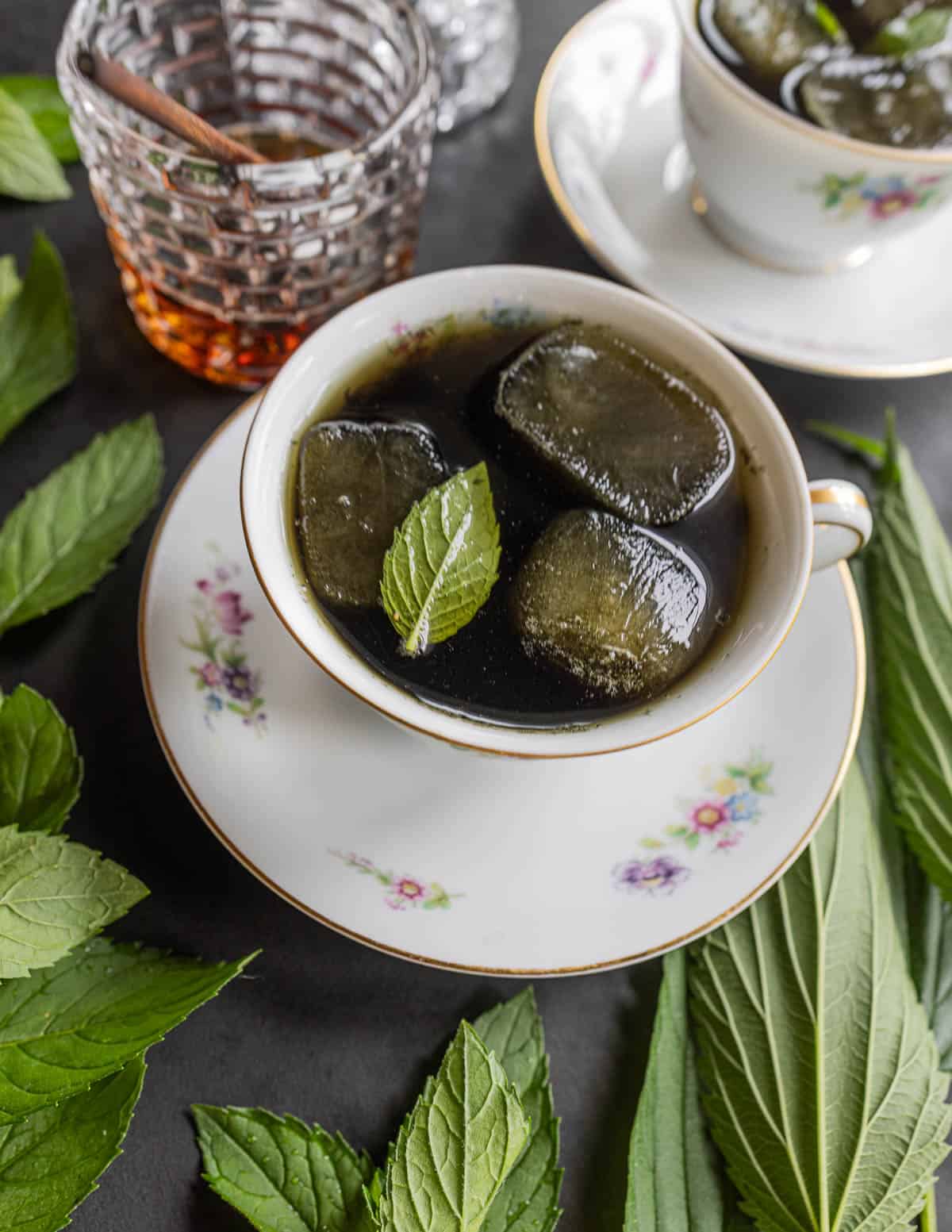 A close up image of two china cups filled with fresh nettle leaf tea garnished with mint, maple syrup and surrounded by fresh wild mint plants and nettle plants. 