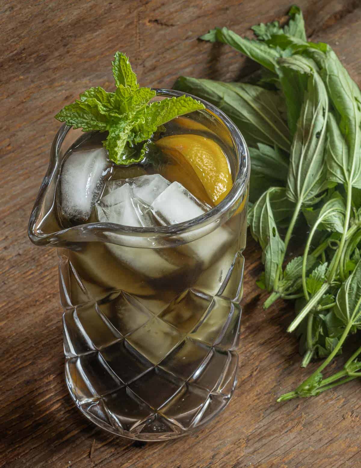 A jar of iced nettle tea with mint and lemon next to fresh stinging nettle plants. 