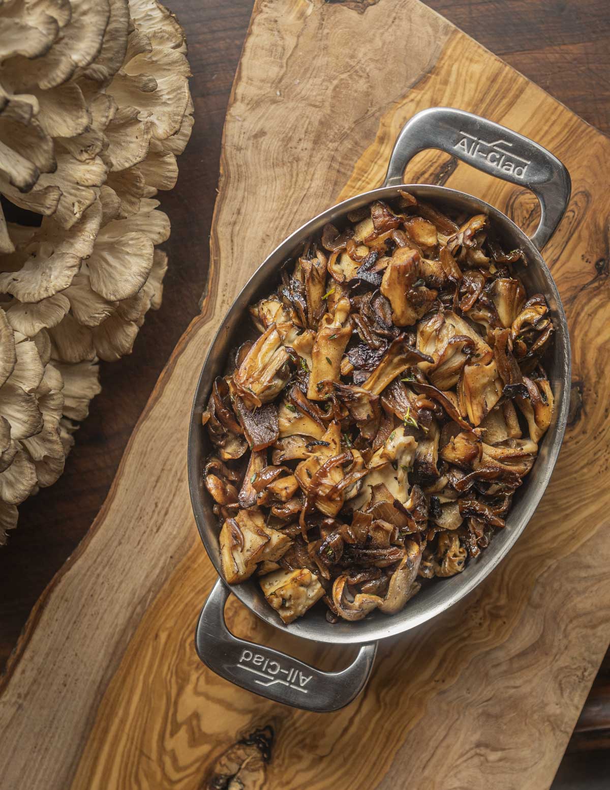 A pan of caramelized mushrooms and onions on a cutting board next to a large hen of the woods mushroom. 