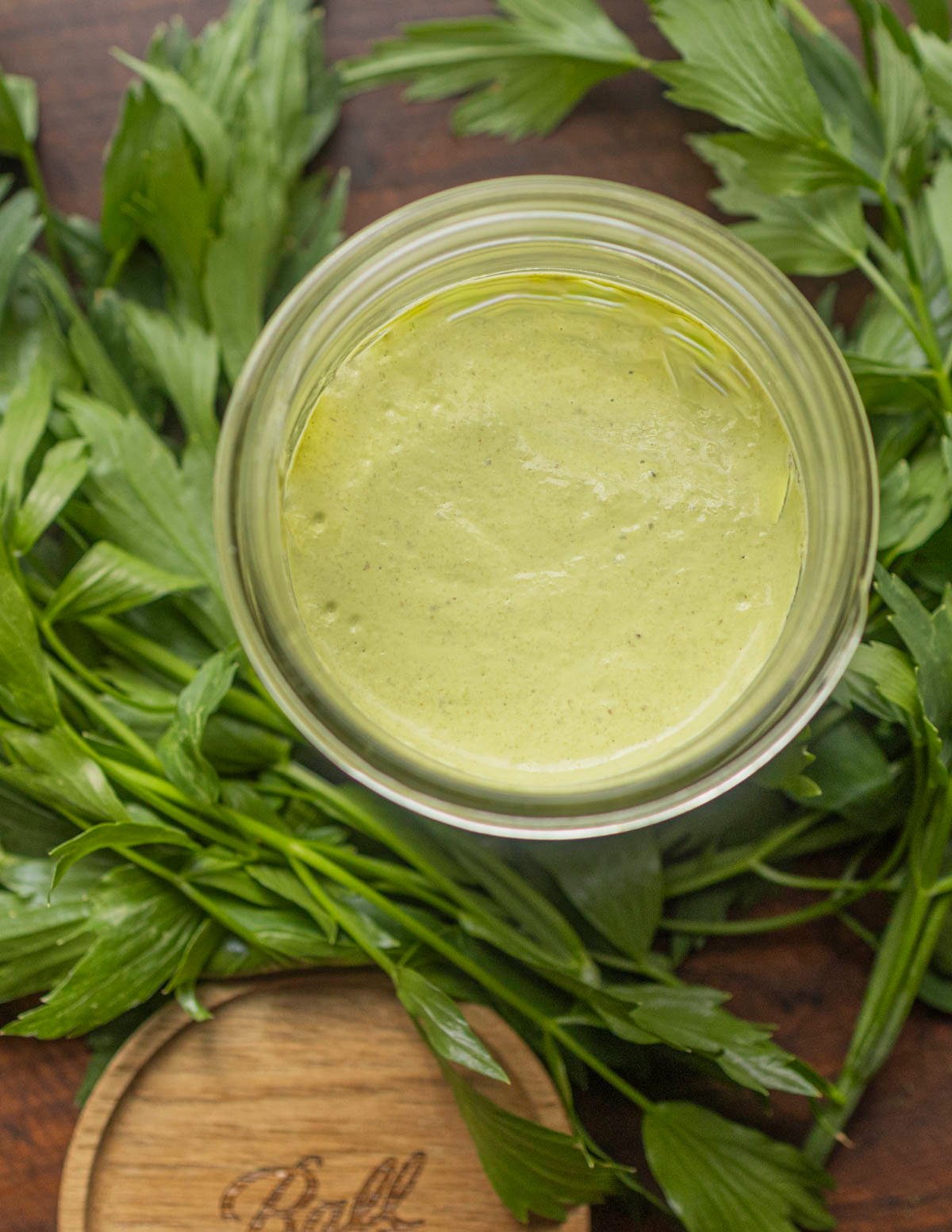 A jar of green lovage herb aioli sauce surrounded by fresh lovage leaves and a wooden mason jar lid. 