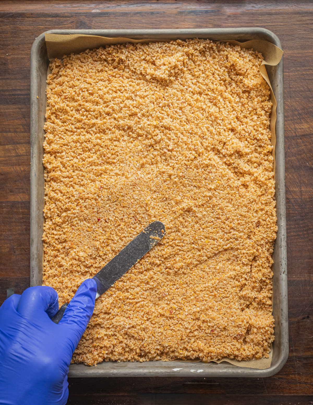 A gloved hand spreading out hot chili pepper salt with a small spatula to dry on a tray. 