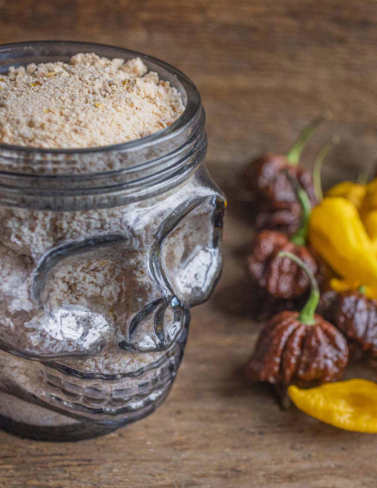 A skull shaped jar filled with spicy hot chili pepper salt next to fresh Carolina reaper peppers and fatalii peppers. 