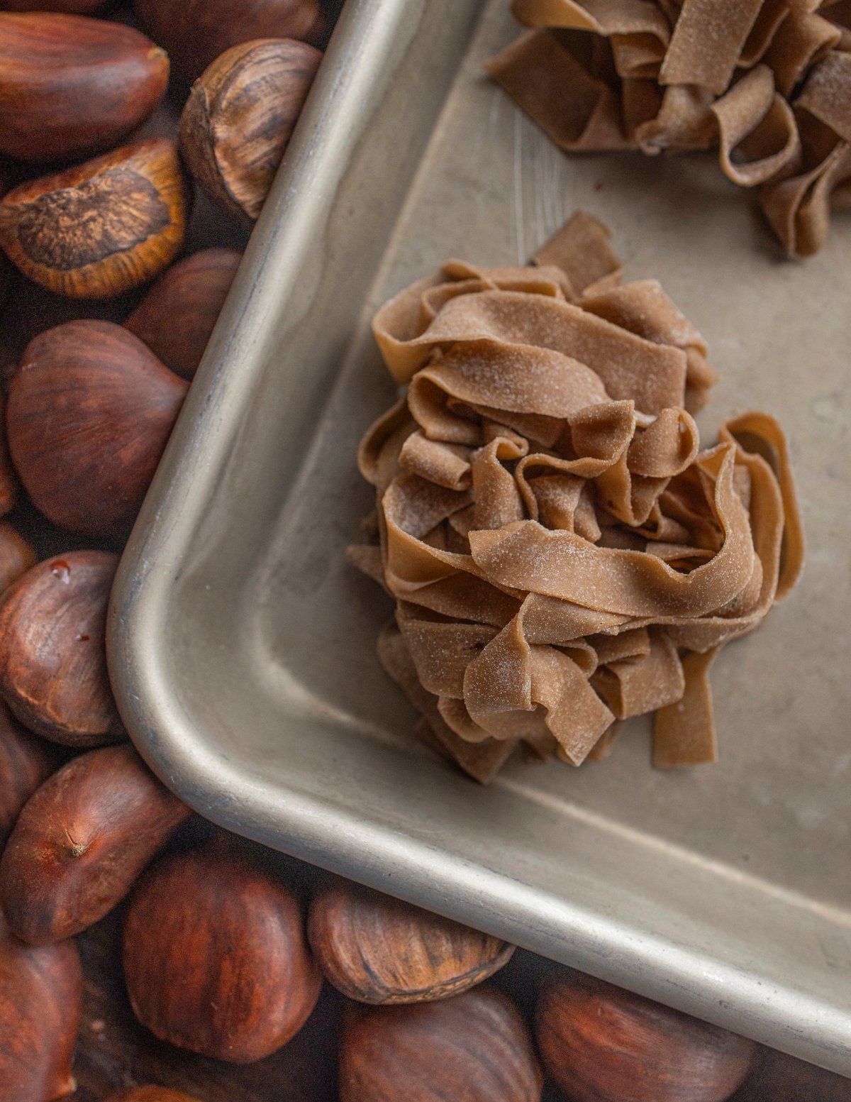 Homemade chestnut flour pasta on a baking tray surrounded by fresh chestnuts. 