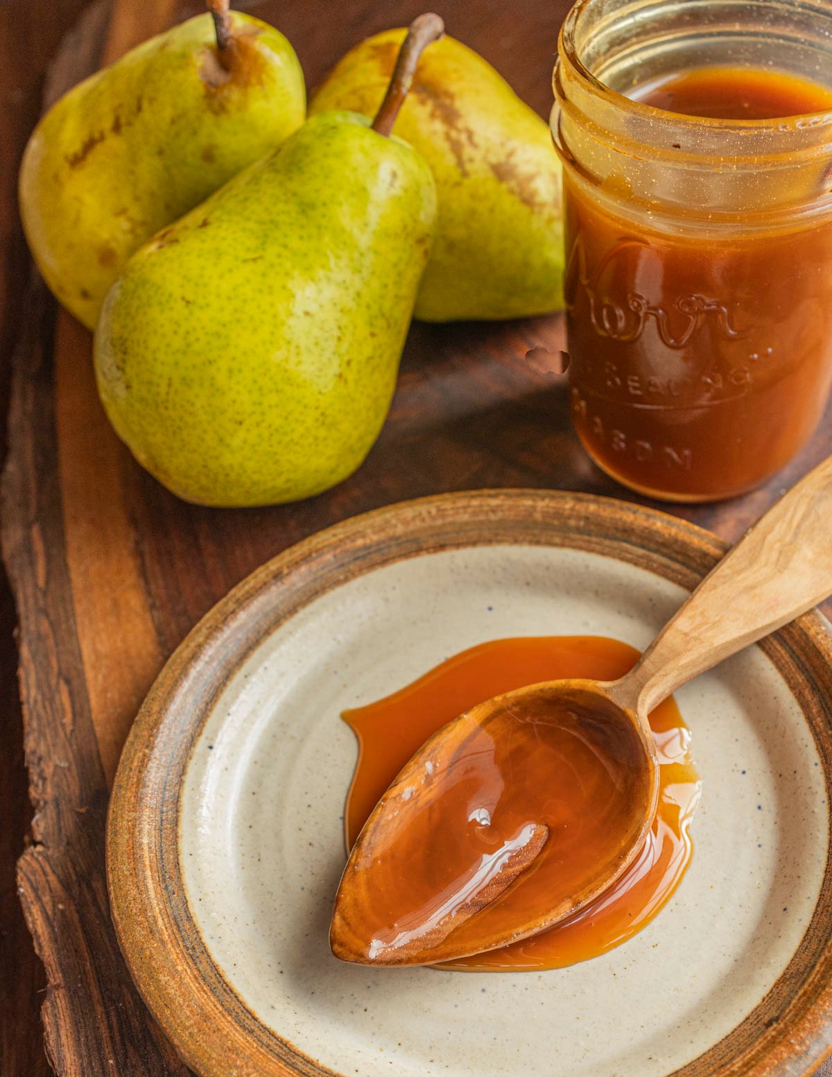 A wooden spoon filled with pear syrup caramel next to fresh pears and a jar of pear syrup. 