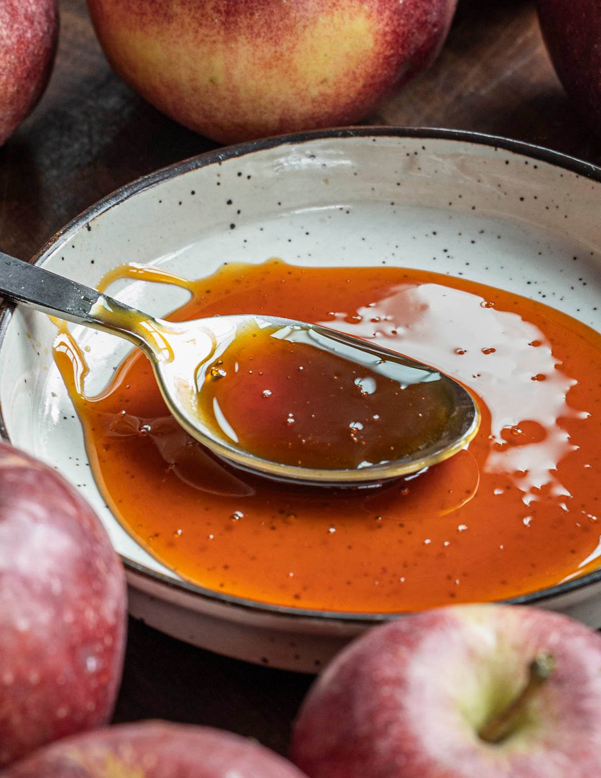 A spoonful of apple molasses on a spoon sitting on a plate surrounded by fresh apples. 