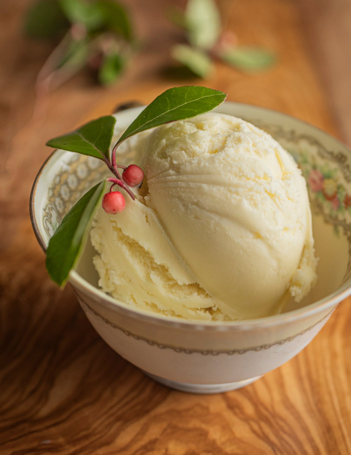 A china cup with a scoop of yellow tea berry ice cream made without food coloring and garnished with teaberries and teaberry leaves. 