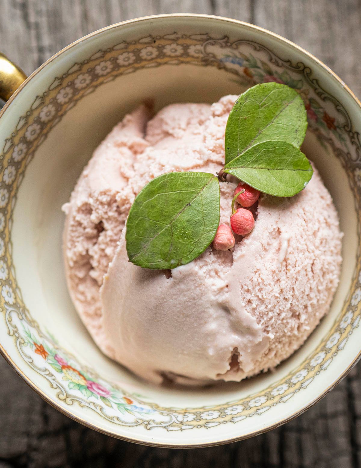 A small china cup filled with a scoop of pink teaberry ice cream garnished with wintergreen leaves and pink wintergreen berries. 