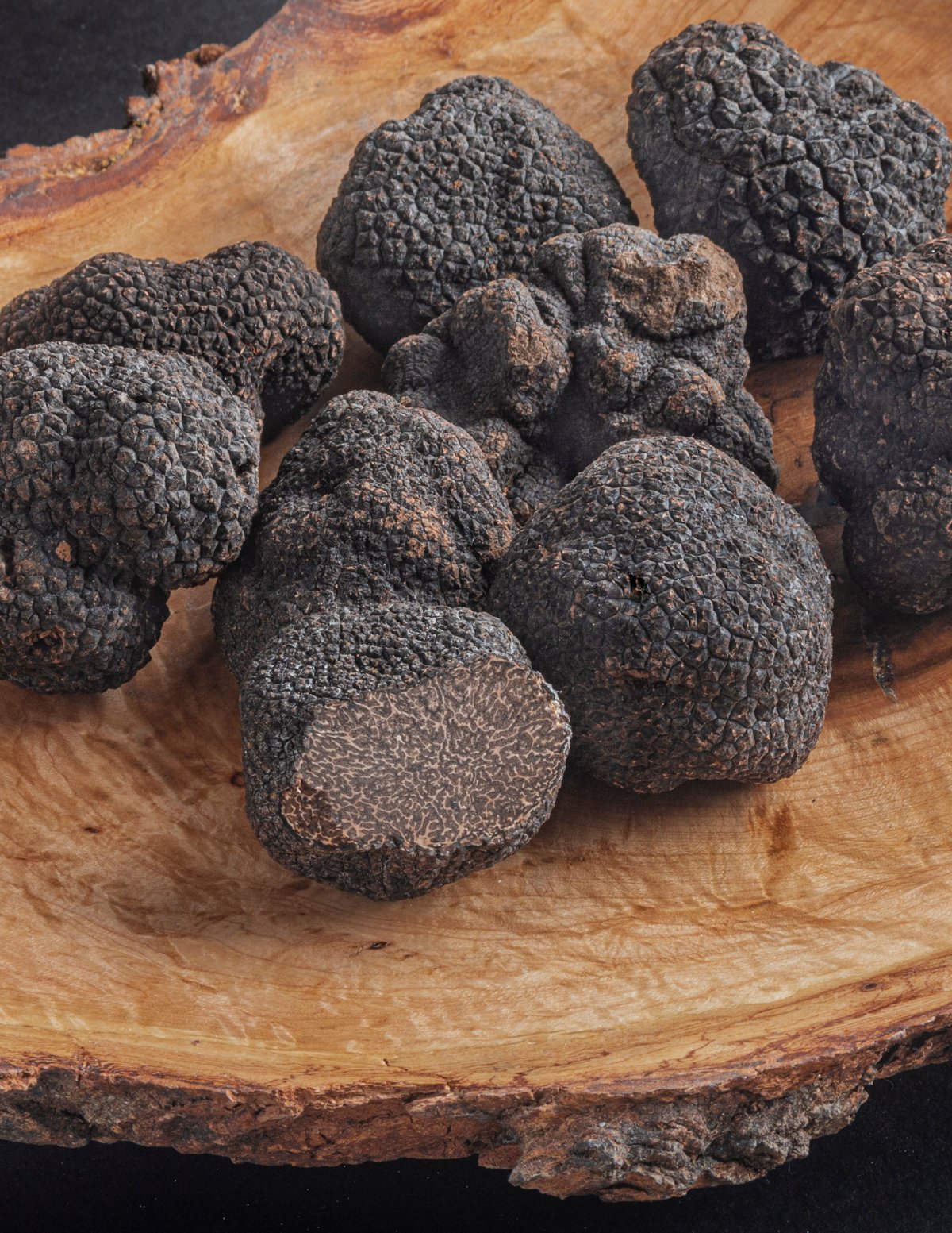 French black Perigord truffles on a wooden plate with one truffle cut open in the front showing the spore bearing surface.